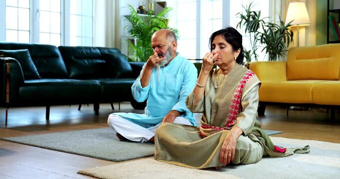 Indian senior couple doing anulom vilom pranayama or yoga at home in the morning sitting with folded legs in modern living room, practicing mindful breathing exercise for health and fitness