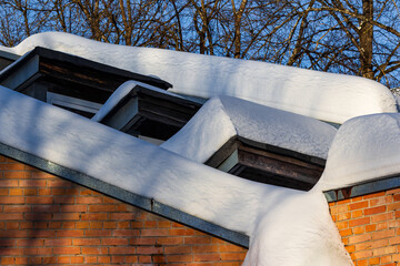 Thick blankets of winter snow drape over a pitched roof with dormer windows, contrasting against a bright blue sky and bare tree branches © PhotoChur
