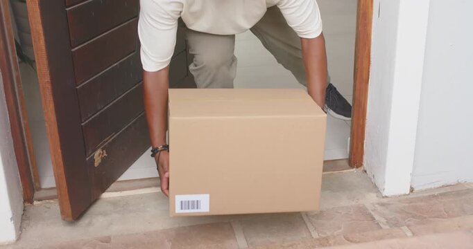 African American man retrieving cardboard box at doorstep, bending, lifting, carrying inside