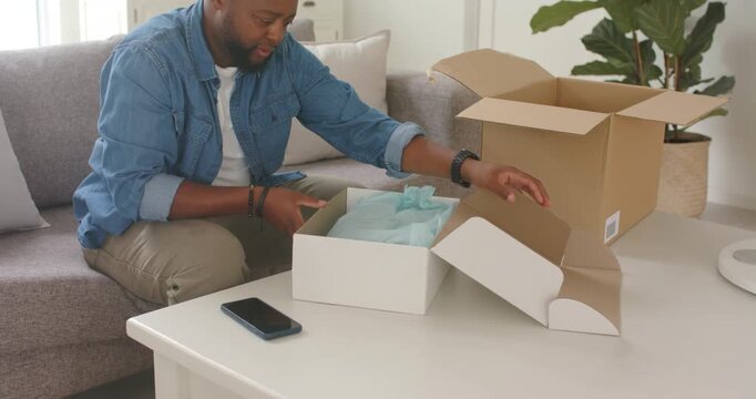African American man placing phone on coffee table, opening white gift box to inspect contents