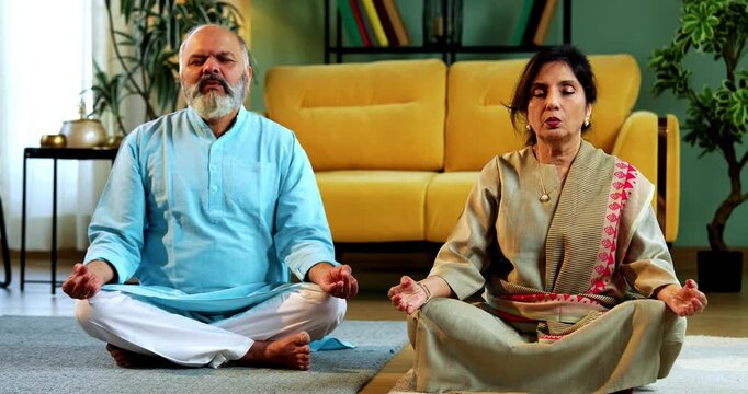 Indian senior couple doing omkar pranayama or yoga at home in the morning sitting with folded legs in modern living room chanting aum, practicing mindful breathing exercise for health