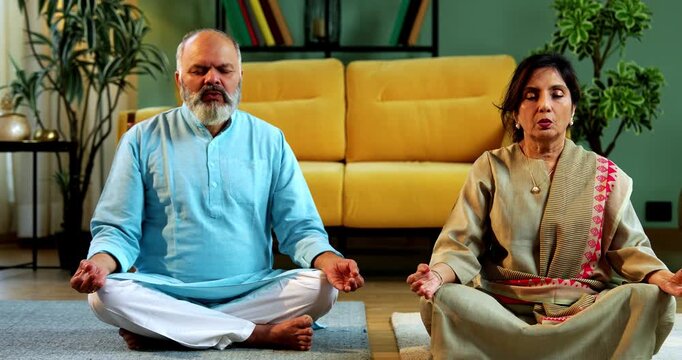 Indian senior couple doing omkar pranayama or yoga at home in the morning sitting with folded legs in modern living room chanting aum, practicing mindful breathing exercise for health