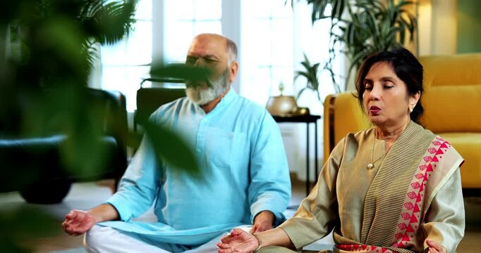 Indian senior couple doing omkar pranayama or yoga at home in the morning sitting with folded legs in modern living room chanting aum, practicing mindful breathing exercise for health