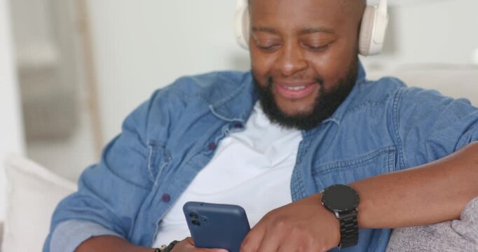 African American man holding blue phone, tapping screen, placing white headphones, smiling on couch