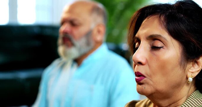 Indian senior couple doing omkar pranayama or yoga at home in the morning sitting with folded legs in modern living room chanting aum, practicing mindful breathing exercise for health