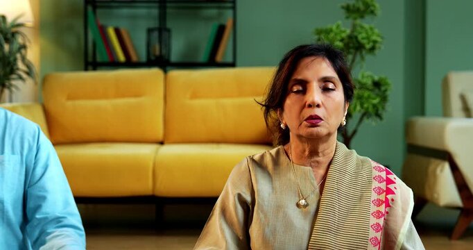 Indian senior couple doing omkar pranayama or yoga at home in the morning sitting with folded legs in modern living room chanting aum, practicing mindful breathing exercise for health