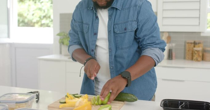 African American man in denim shirt slicing cucumber, pepper on board in kitchen preparing meals
