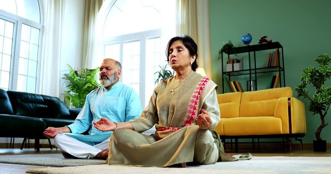Indian senior couple doing omkar pranayama or yoga at home in the morning sitting with folded legs in modern living room chanting aum, practicing mindful breathing exercise for health