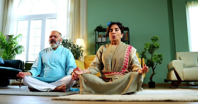 Indian senior couple doing omkar pranayama or yoga at home in the morning sitting with folded legs in modern living room chanting aum, practicing mindful breathing exercise for health