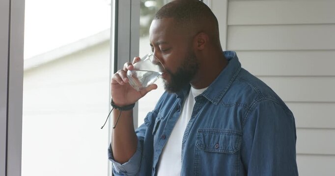 Man 30s standing at backlit window, wearing denim-shirt, sipping half-full glass, finding resolve