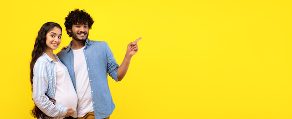 A couple stands together in a bright setting. The man points to the side while the woman smiles and holds her belly. Both appear excited about their news as they pose against a yellow backdrop. © Prostock-studio