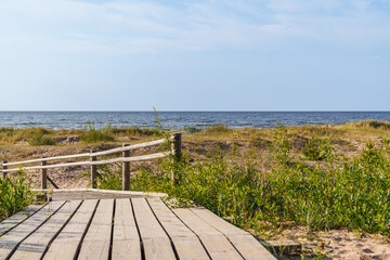 Fototapeta premium A wooden boardwalk leads to the Baltic sea in Latvia. The beach is covered in grass and there is a clear blue sky above