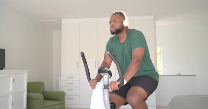 African man fitting headphones pedaling bike in bedroom wiping with towel checking watch for cardio