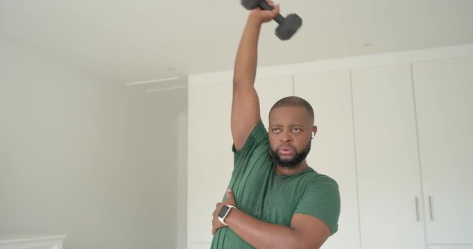 Adult African man holding dumbbell overhead, working triceps for strength at home in green tee