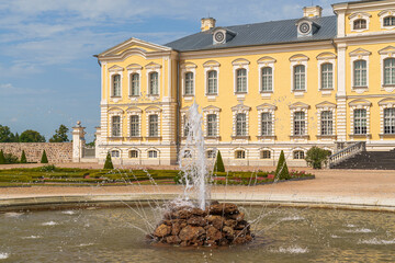 Grand yellow Rundale palace with classical architecture. Fountain jets water in center of stone pond. Symmetrical gardens frame the elegant structure. Sunny sky enhances the scene's calm elegance