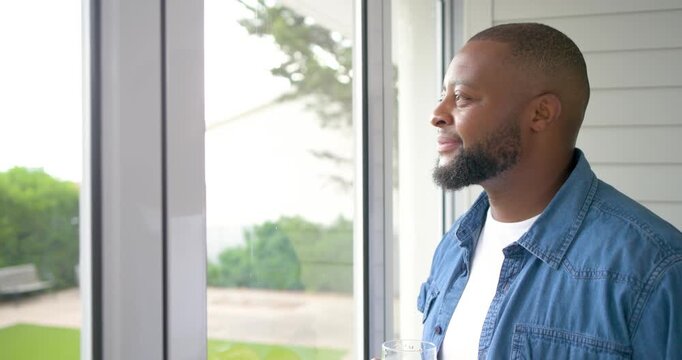 African American man at home by sliding door holding glass, viewing yard and reflecting, copy space