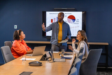 Diverse team wearing business attire presenting around meeting table with wall display using tablet