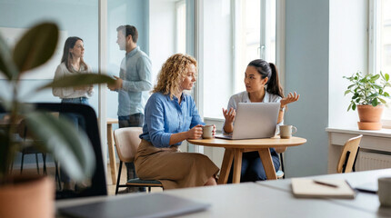 Fototapeta premium Two women collaborating with a laptop at a small round table in a modern, bright office setting while colleagues talk in the background