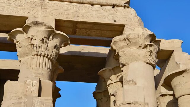 Kom Ombo temple columns with composite capitals and hieroglyphic carvings viewed from below against blue sky in Egypt, upward perspective of ancient sandstone architecture.