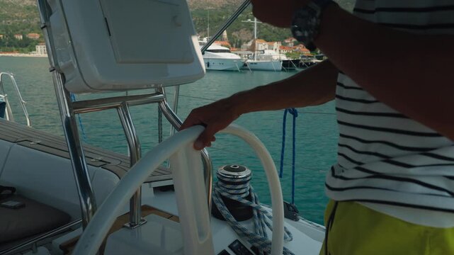 Skipper At Helm Steering Yacht Cockpit, Hand On Wheel And Striped Shirt Visible, View Toward Harbor And Passing Motorboats With Relaxed Summer Sailing Vibe