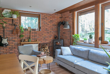 Naklejka premium Rural cottage with cozy design, windows to the forest, fireplace, sofa, armchair and red brick wall. Interior of living room with design indoor architecture.