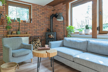 Naklejka premium Rural cottage with cozy design, windows to the forest, fireplace, sofa, armchair and red brick wall. Interior of living room with design indoor architecture. 