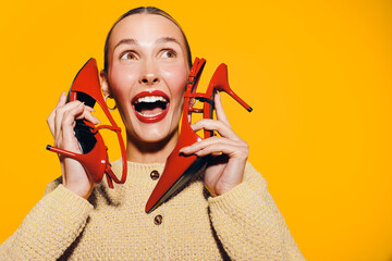 Happy woman in beige sweater holds red high heels near face, smiling widely on bright yellow...