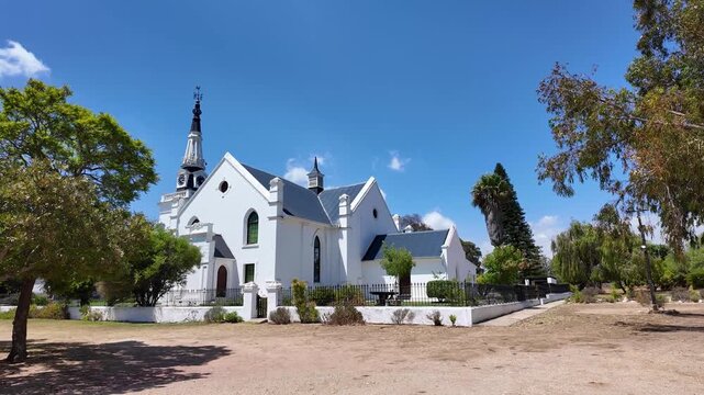 Hopefield South Africa. 10.02.2026. A panoramic view of a classic Dutch Reform church building with a tall spire, surrounded by green trees and a dusty ground on a sunny day in Hopefield South Africa.
