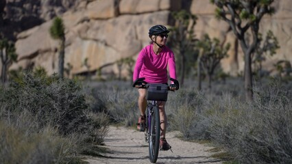 Fototapeta premium A mature woman rides her bike on a dirt road in Joshua Tree National Park, surrounded by unique rock formations and plants