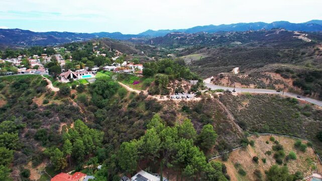 drone footage flying over an upscale hillside community nestled in rolling green hills. The scenic landscape features luxury residential homes with swimming pools, winding mountain roads.