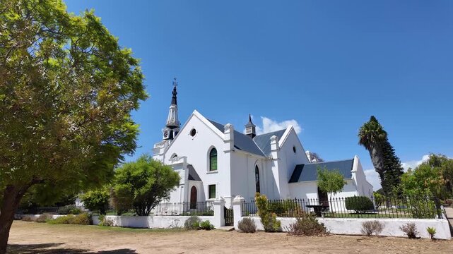Hopefield South Africa. 10.02.2026. A panoramic view of a classic Dutch Reform church building with a tall spire, surrounded by green trees and a dusty ground on a sunny day in Hopefield South Africa.