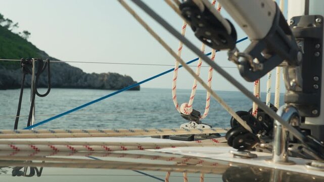 Closeup Rigging Pulleys Ropes Foredeck Crew Member Adjusting Foresail Hardware, Blue Sea And Rocky Coastline In Background, Brass Fittings, Taut Lines, Nautical Work