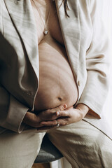Stylish pregnant woman hugging baby bump in window light, close up. Happy Mothers day! Healthy pregnancy and fertility concept. Fashionable woman in suit holding pregnant belly