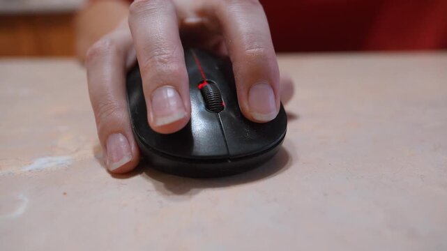 Close-up view of a woman's hand using a black wireless computer mouse on a desk, clicking the buttons and scrolling the wheel while working, studying or browsing the internet online