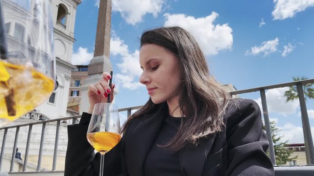 Stylish brunette businesswoman drinking aperol spritz orange cocktail while sitting in restaurant at Spanish steps in Rome with background view of obelisk and church Trinit&agrave; dei Monti, cinematic