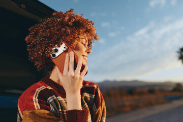 Curly woman smiling outdoors, holding smartphone to ear, wearing colorful sweater, enjoying sunny...