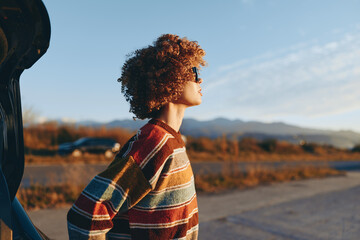 Woman with curly hair wearing a rainbow sweater stands outdoors near a car, looking into the distance with a calm smile, soft sunlight, and mountains in the background. © SHOTPRIME STUDIO