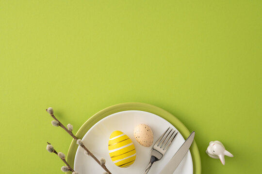 Happy Easter tabletop composition featuring a painted yellow egg, speckled egg, cutlery and a pussy willow branch beside a small ceramic bunny on a bright green background