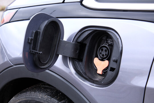Close-up of open charging port on silver electric car. Gray metallic body panel, black plastic flap hinged open, orange Type 2 connector socket with three-phase pins and central pilot, small latch mec