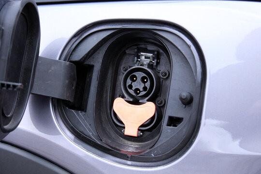 Close-up of open charging port on silver electric car. Gray metallic body panel, black plastic flap hinged open, orange Type 2 connector socket with three-phase pins and central pilot, small latch mec