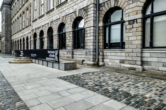 Stone facade of the Old Library at Trinity College Dublin, Ireland, featuring arched windows and "The Book of Kells Experience" signage.