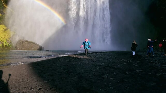 Running happy female tourist on visit of the Skogafoss waterfall flowing from cliff in Iceland, a famous place and travel destination, slow motion