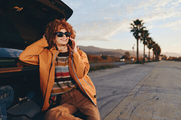 Smiling woman with curly hair wearing a rainbow sweater and orange jacket sits by the open car...