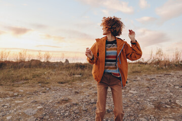 Woman with curly hair wearing rainbow sweater and orange jacket stands outdoors on rocky ground during sunset smiling and enjoying lifestyle with positive mood and casual style. © SHOTPRIME STUDIO