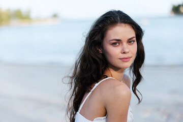 Portrait of a young woman with dark hair on a sandy beach, wearing a light top, gazing softly at...