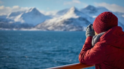 Fototapeta premium Traveler in red winter gear photographing snowy mountains over blue ocean water from a ship deck