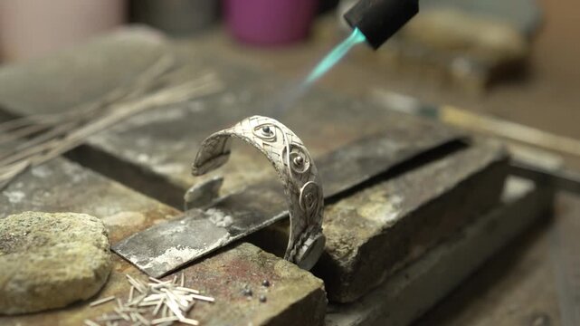 Close-up of a silversmith using a torch to solder a detailed ornate bracelet on a workbench.