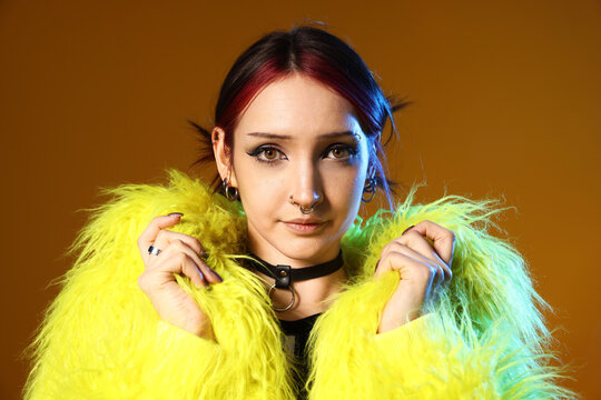 Portrait of beautiful young woman with piercings and choker in fur coat in color light on brown background