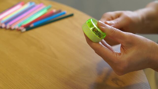 A close-up shot of a woman's hands using a small green manual sharpener to sharpen an orange pencil over a wooden table