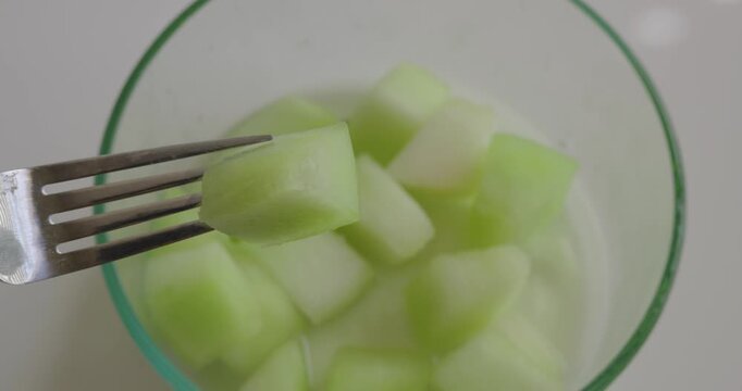 A honeydew melon chunk held on a fork above a glass bowl